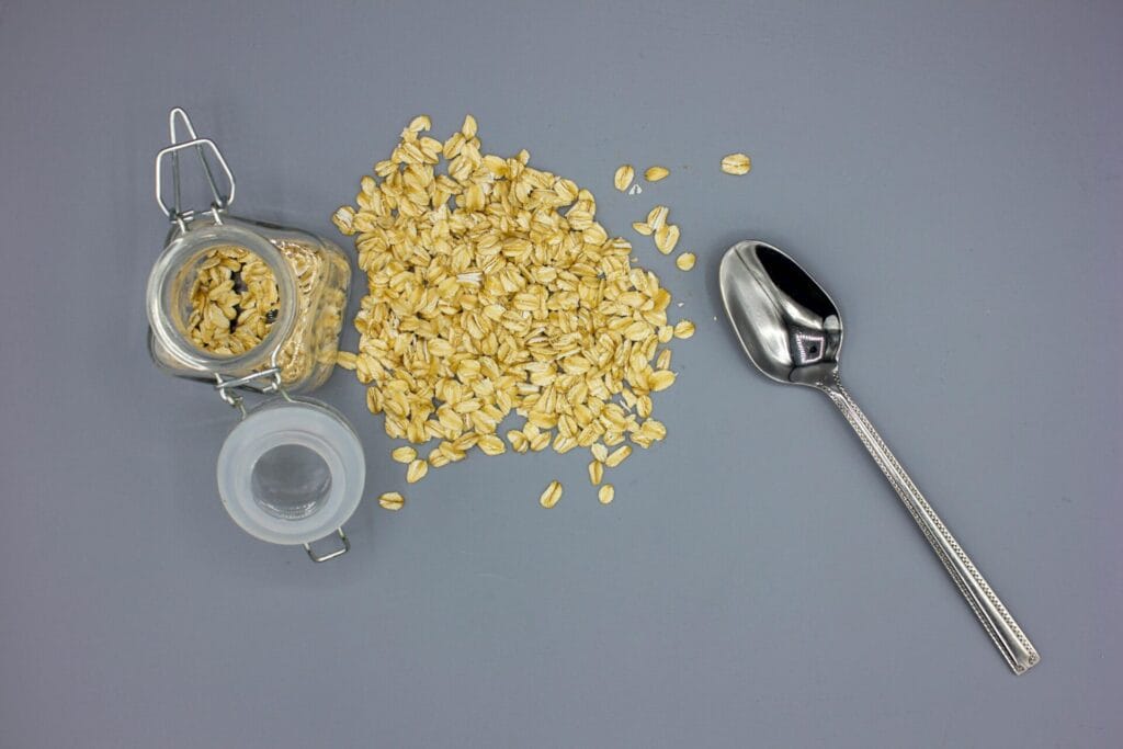 Overhead shot of scattered oats with a glass jar and silver spoon on a gray surface, perfect for breakfast themes.