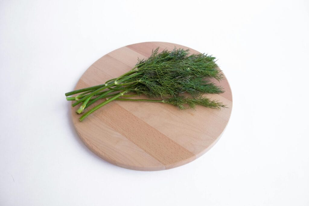 A close-up of fresh dill herb resting on a round wooden chopping board against a white background.