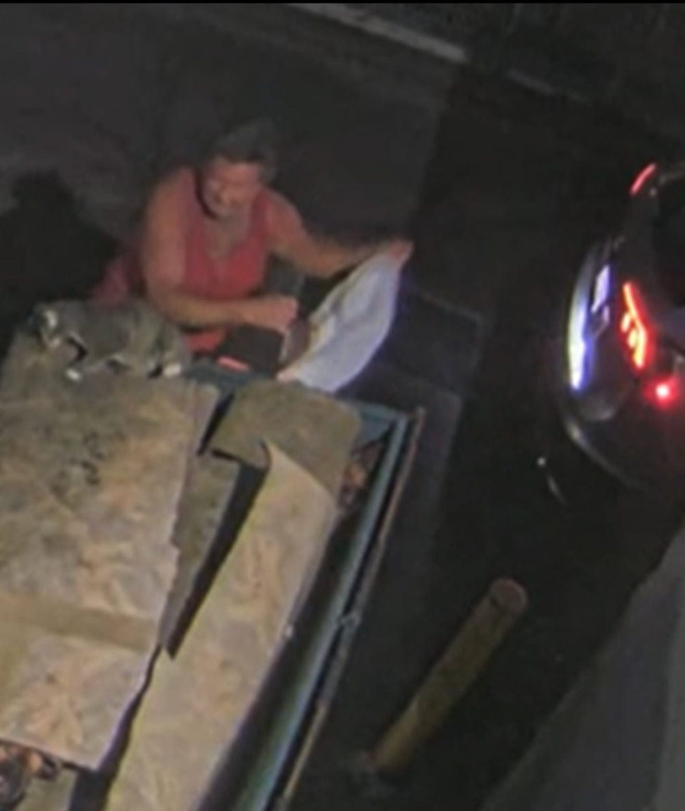 Nighttime surveillance footage shows a man extending a white bag over the edge of a dumpster.